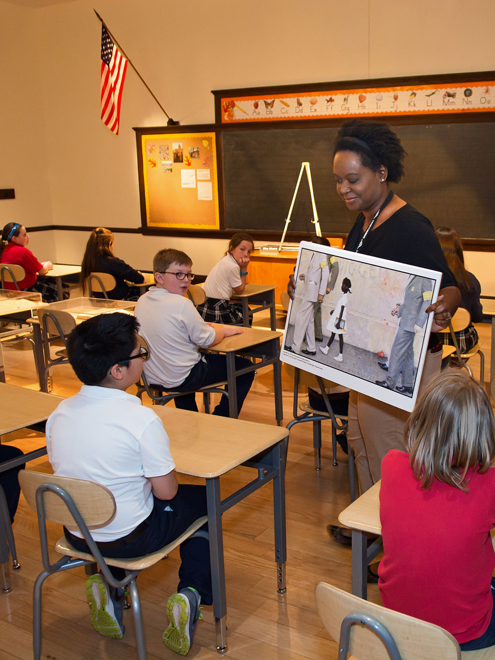 Children sitting at classroom desks looking at a copy of Norman Rockwell's painting The Problem We All Live With.